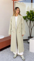 Front full view of woman wearing Sage Green double-breasted maxi trench coat with white wide-leg trousers